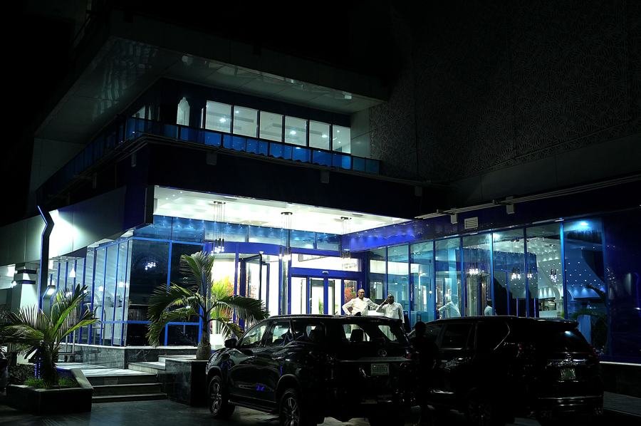 Night shot of blue-lit glass entrance facade with canopy, palm plants and black SUVs parked in front; people at the doors.