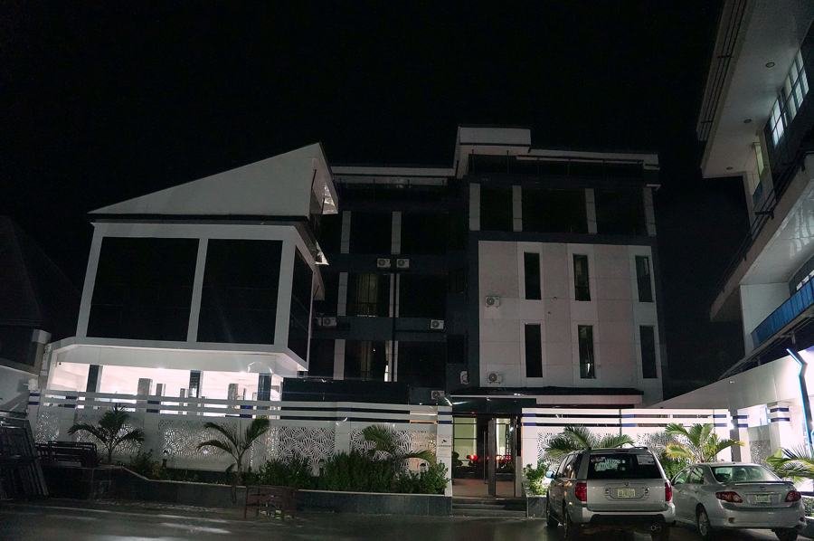 Dark night view of the white multi-storey building from the street over the perimeter wall, angular glazed pavilion at left, parked cars; underexposed.