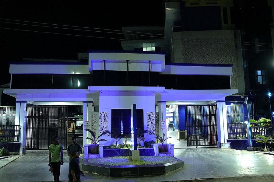 Front gatehouse/portico bathed in royal-blue-violet uplighting with central fountain planter, symmetric composition, two bystanders at left; the strongest and most on-brand night shot of the