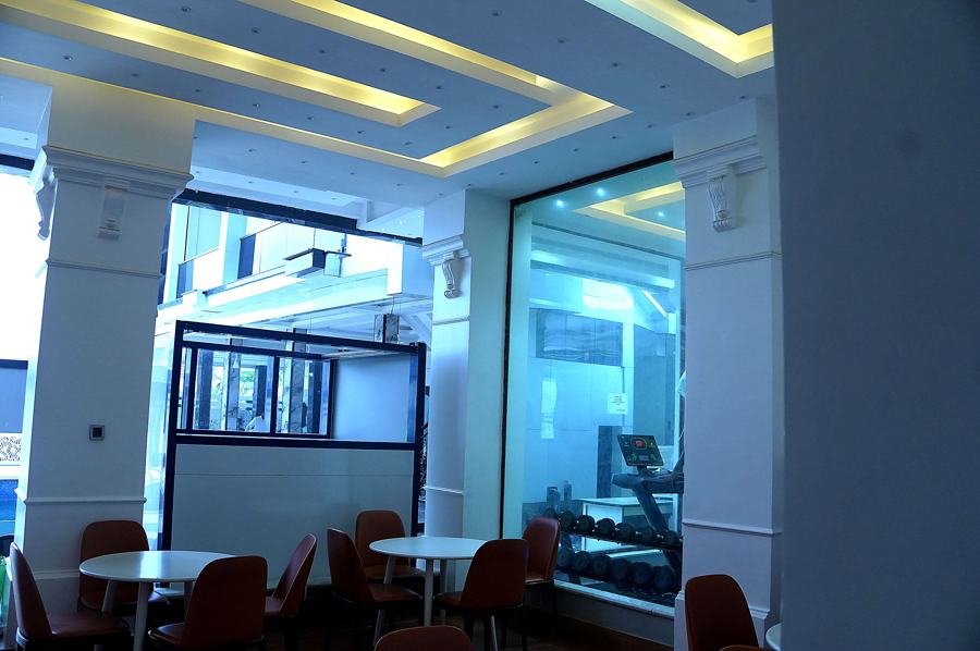 Cafe/breakfast seating area: round white tables with reddish-brown chairs, coffered ceiling with warm cove lighting, glass wall looking into the gym