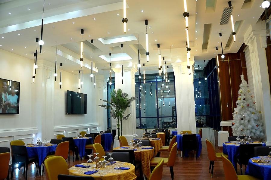 Wide dining-room view: tall white ceiling with linear drop pendants, tall windows, blue/gold dressed tables, yellow chairs, white Christmas tree