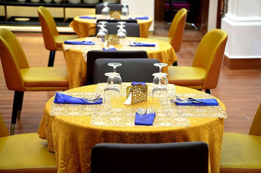 Round table with gold cloth, blue napkins, mustard chairs on wood floor; second dressed table and buffet line behind