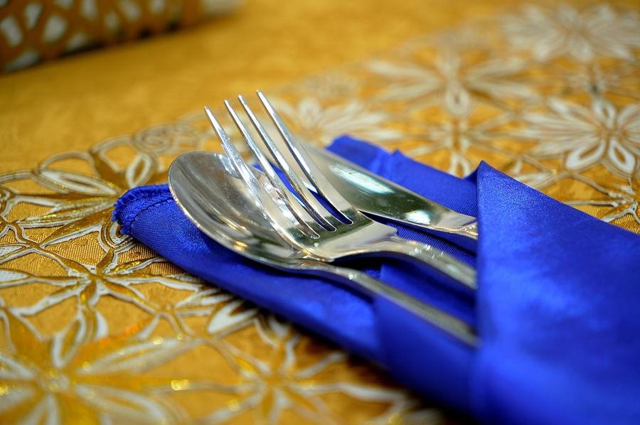Macro of silver fork and spoon on royal blue napkin over gold embroidered tablecloth; very on-brand blue/gold detail