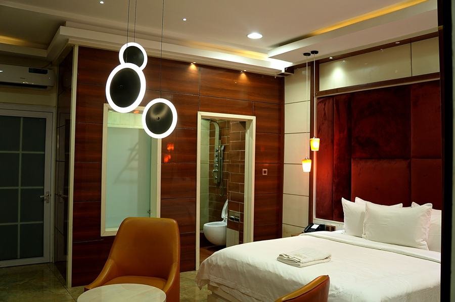 Wood-panel room, maroon velvet padded headboard wall, white bed, orange armchair, ring pendant lights, frosted-glass bathroom partition